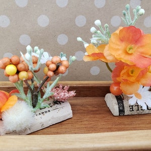 May include: Two small floral arrangements in orange and white, each with a single flower on a cork base. The cork bases have text on them, one says "Bottle" and the other says "Leyards".