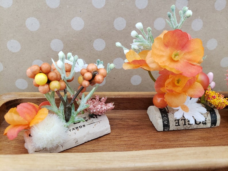 May include: Two small floral arrangements in orange and white, each with a single flower on a cork base. The cork bases have text on them, one says "Bottle" and the other says "Leyards".