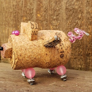 May include: A whimsical cork pig figurine with pink beads for eyes and legs, a pink and silver wire tail, and a silver butterfly wing. The cork is labeled "51-18".