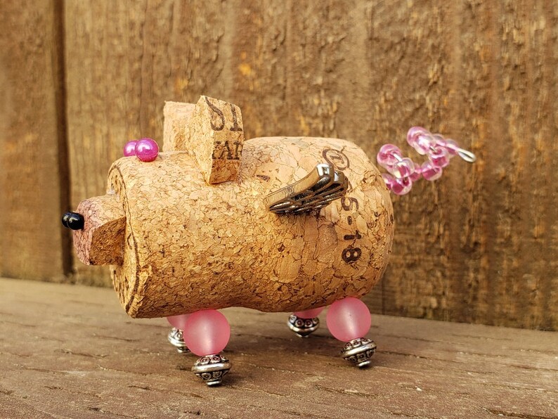 May include: A whimsical cork pig figurine with pink beads for eyes and legs, a pink and silver wire tail, and a silver butterfly wing. The cork is labeled "51-18".