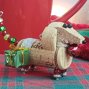 May include: A Christmas ornament shaped like a dachshund made from a wine cork. The ornament is decorated with green and red beads, a small green gift box, and a red bell. The cork is labeled "Diageo" and "Ch&acirc;teau".