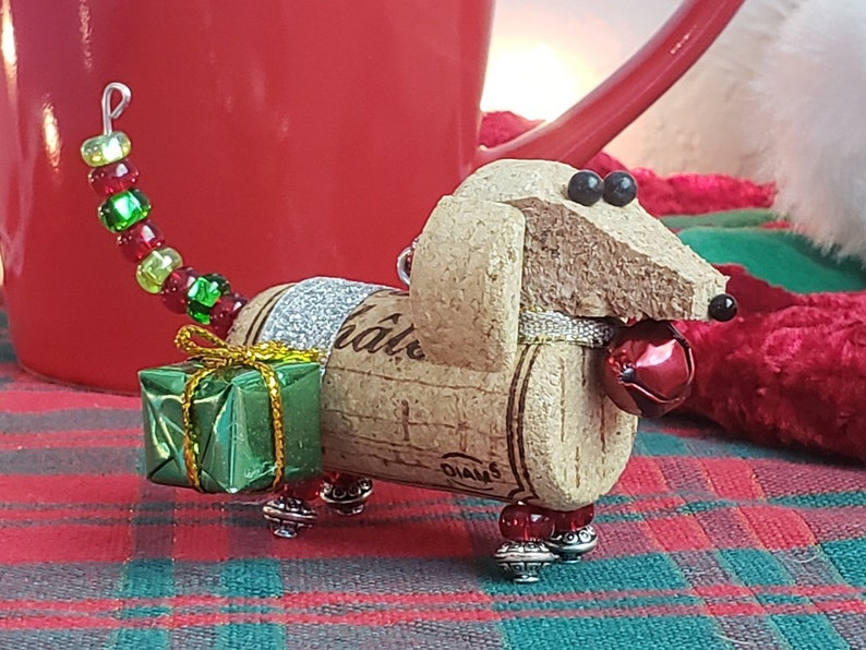 May include: A Christmas ornament shaped like a dachshund made from a wine cork. The ornament is decorated with green and red beads, a small green gift box, and a red bell. The cork is labeled "Diageo" and "Ch&acirc;teau".