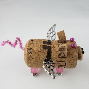 May include: A pink and silver winged pig figurine made from a wine cork. The pig has pink beads for legs and a pink beaded tail. The wings are silver and have a delicate design.