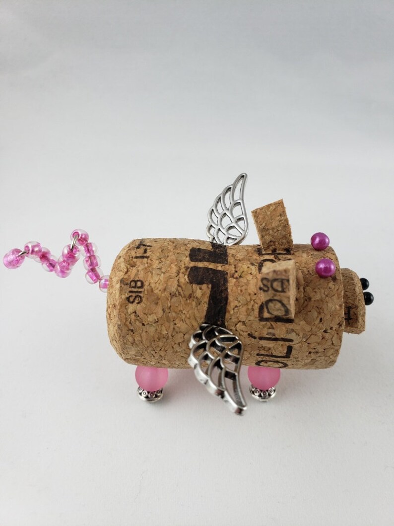 May include: A pink and silver winged pig figurine made from a wine cork. The pig has pink beads for legs and a pink beaded tail. The wings are silver and have a delicate design.