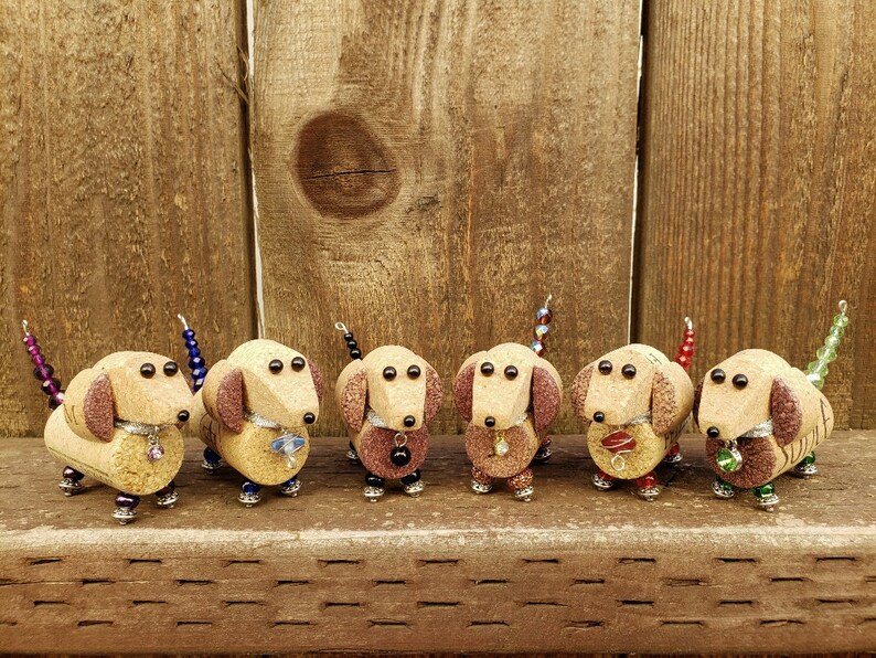 May include: Six miniature dachshund figurines made from wine corks and decorated with beads. The dachshunds are brown and have black eyes. Each dachshund has a different colored beaded tail.