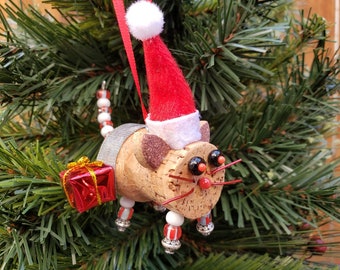 May include: A cork cat ornament wearing a red Santa hat and holding a small red gift box. The cat is decorated with silver and white beads and has black eyes.