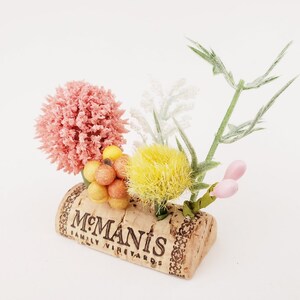 May include: A small floral arrangement in a cork with the text "McManis Family Vineyards". The arrangement includes pink, yellow, and white flowers, as well as small orange and yellow berries.