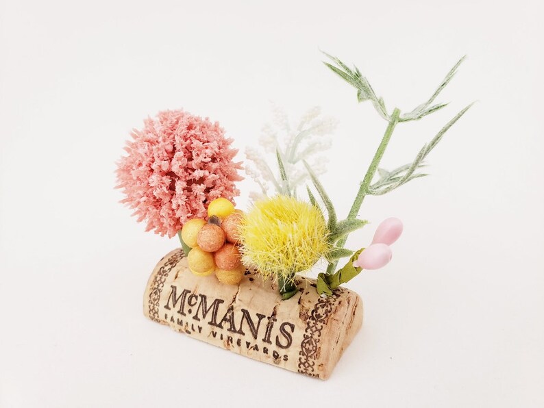 May include: A small floral arrangement in a cork with the text "McManis Family Vineyards". The arrangement includes pink, yellow, and white flowers, as well as small orange and yellow berries.