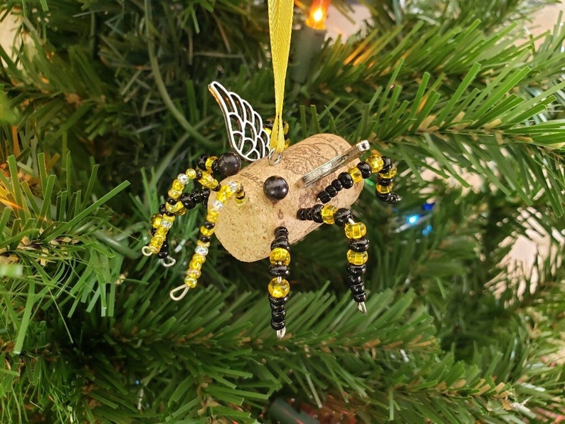 Puede incluir: Un adorno de abeja de corcho con cuentas negras y amarillas, alas plateadas y una antena de alambre. El adorno est&aacute; colgado de un &aacute;rbol de Navidad verde.