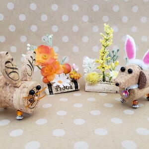 May include: Two cork bunny and dachshund figurines with floral accents. The bunny is brown with black eyes and a gold wire nose. The dachshund is brown with black eyes and a pink bunny ear headband. Both figurines are on a brown background with white polka dots.