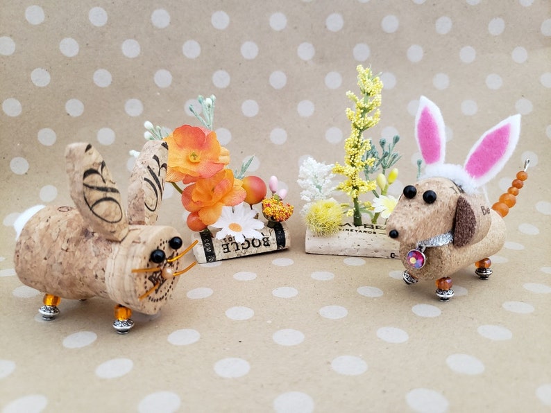 May include: Two cork bunny and dachshund figurines with floral accents. The bunny is brown with black eyes and a gold wire nose. The dachshund is brown with black eyes and a pink bunny ear headband. Both figurines are on a brown background with white polka dots.