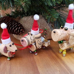 May include: Three cork dog ornaments with red Santa hats and beaded details. Each dog is holding a small gift box. The ornaments are on a wooden surface.
