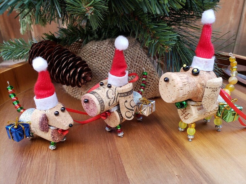 May include: Three cork dog ornaments with red Santa hats and beaded details. Each dog is holding a small gift box. The ornaments are on a wooden surface.