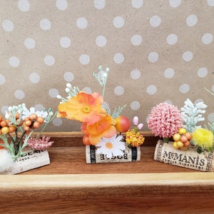 May include: Three small floral arrangements in cork wine bottle stoppers. The arrangements feature orange, yellow, pink, and white flowers with green leaves. The stoppers are labeled "El Loco", "M.Manis Family Vineyards", and "Dear Yards".