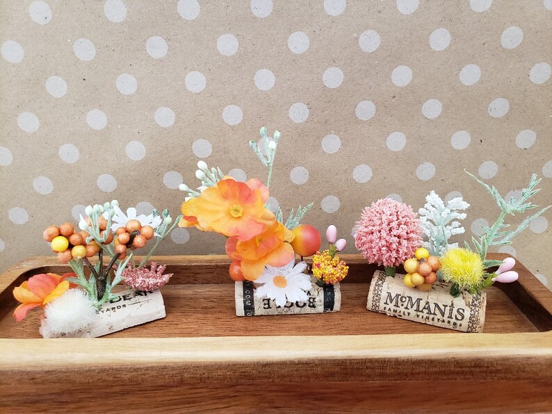 May include: Three small floral arrangements in cork wine bottle stoppers. The arrangements feature orange, yellow, pink, and white flowers with green leaves. The stoppers are labeled "El Loco", "M.Manis Family Vineyards", and "Dear Yards".