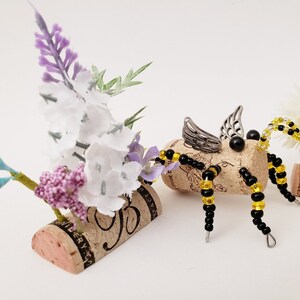 Puede incluir: Una abeja caprichosa hecha de un corcho de vino, cuentas negras y amarillas y alas plateadas. La abeja est&aacute; posada sobre una base de corcho con un arreglo floral de flores blancas y moradas.