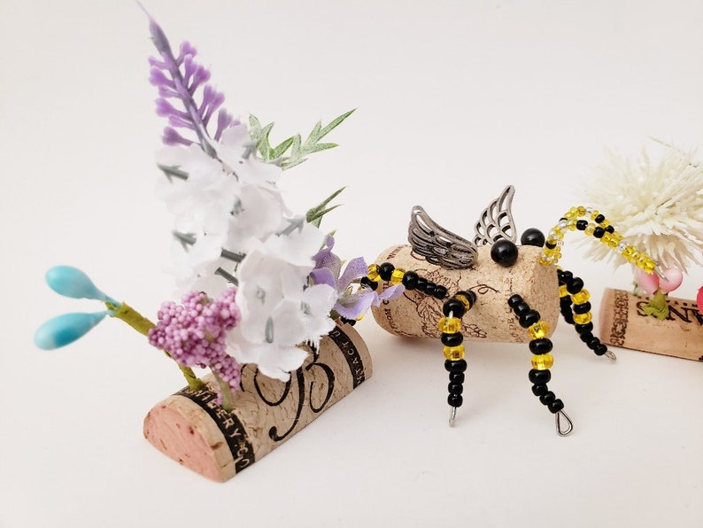 Puede incluir: Una abeja caprichosa hecha de un corcho de vino, cuentas negras y amarillas y alas plateadas. La abeja est&aacute; posada sobre una base de corcho con un arreglo floral de flores blancas y moradas.