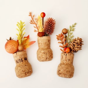 May include: Three decorative corks with autumn-themed floral arrangements. Each cork features a different arrangement of fall foliage, including berries, leaves, and pinecones, in shades of orange, yellow, and brown. The corks are arranged on a white background.