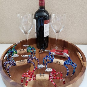 May include: Five colorful beaded crab bottle stoppers on a wooden tray with two wine glasses and a bottle of red wine. The crabs are made with glass beads and have a cork body. The tray is round and has a brown finish.