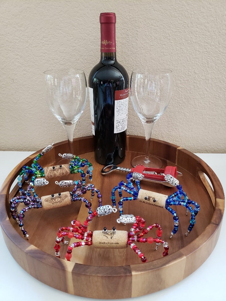 May include: Five colorful beaded crab bottle stoppers on a wooden tray with two wine glasses and a bottle of red wine. The crabs are made with glass beads and have a cork body. The tray is round and has a brown finish.