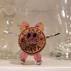 May include: A small pig figurine made from a wine cork with pink beads for legs and pink and purple beads for ears. The cork has the word "CAVA" printed on it.
