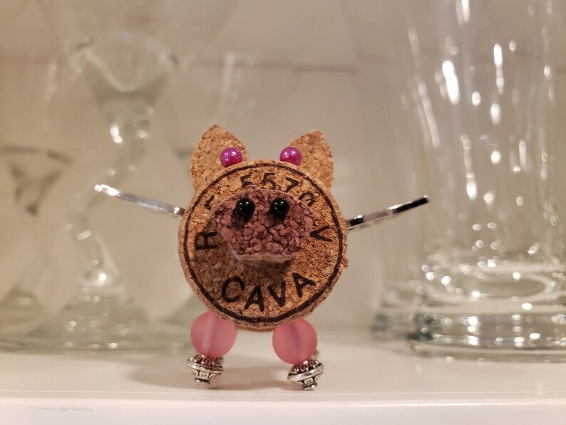 May include: A small pig figurine made from a wine cork with pink beads for legs and pink and purple beads for ears. The cork has the word "CAVA" printed on it.