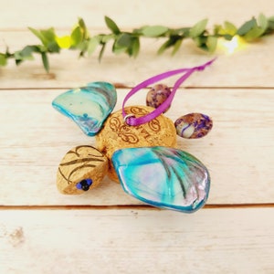 May include: A handmade ornament shaped like a turtle with iridescent blue wings, a cork body, and purple beads. The ornament is strung with a purple ribbon for hanging.