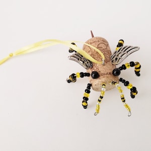 Puede incluir: Un adorno de abeja de corcho con cuentas negras y amarillas, alas plateadas y una cinta amarilla para colgar.