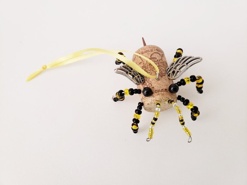 Puede incluir: Un adorno de abeja de corcho con cuentas negras y amarillas, alas plateadas y una cinta amarilla para colgar.