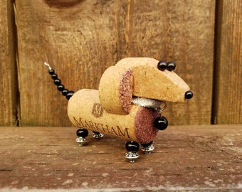 May include: A dachshund-shaped figurine made from wine corks, with black beads for eyes and a silver collar with a black bead charm. The figurine is standing on a wooden surface.