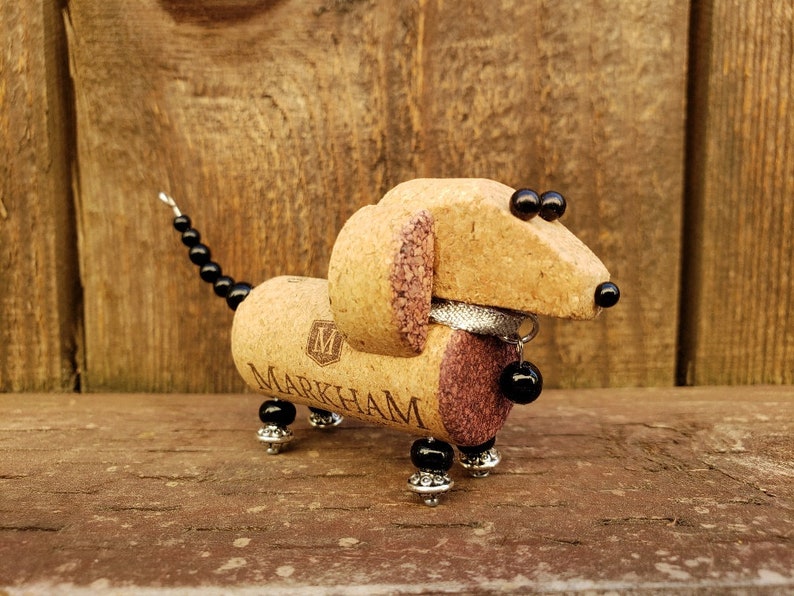 May include: A dachshund-shaped figurine made from wine corks, with black beads for eyes and a silver collar with a black bead charm. The figurine is standing on a wooden surface.