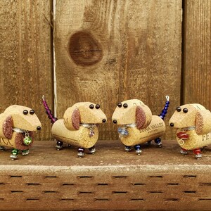 May include: Four miniature dachshund figurines made from wine corks, each with a different colored tail and beads. The dachshunds are standing on a wooden surface.