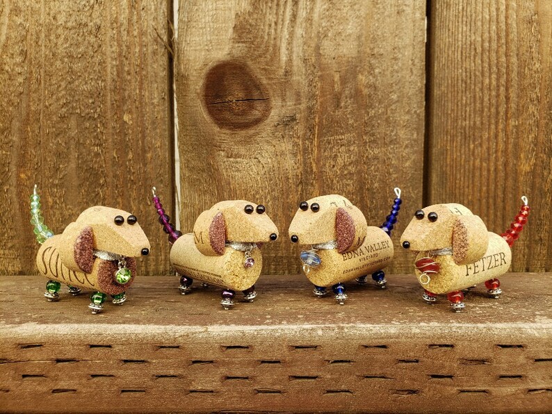 May include: Four miniature dachshund figurines made from wine corks, each with a different colored tail and beads. The dachshunds are standing on a wooden surface.