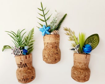 May include: Three cork bottle stoppers decorated with miniature sea-themed items. The stoppers are topped with green and blue plants, a blue starfish, a seashell, a silver anchor, a blue whale tail, and a blue glass bead.