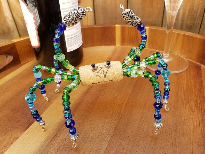 May include: A beaded wine bottle stopper shaped like a crab with green, blue, and clear beads. The crab has silver accents and is sitting on a wooden surface.