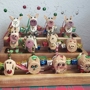 May include: A tiered wooden display stand with 12 handmade reindeer and cat ornaments made from wine corks. The ornaments are decorated with beads, bells, and ribbons. The reindeer have red noses and antlers made from wire. The cats have whiskers and tails made from wire.