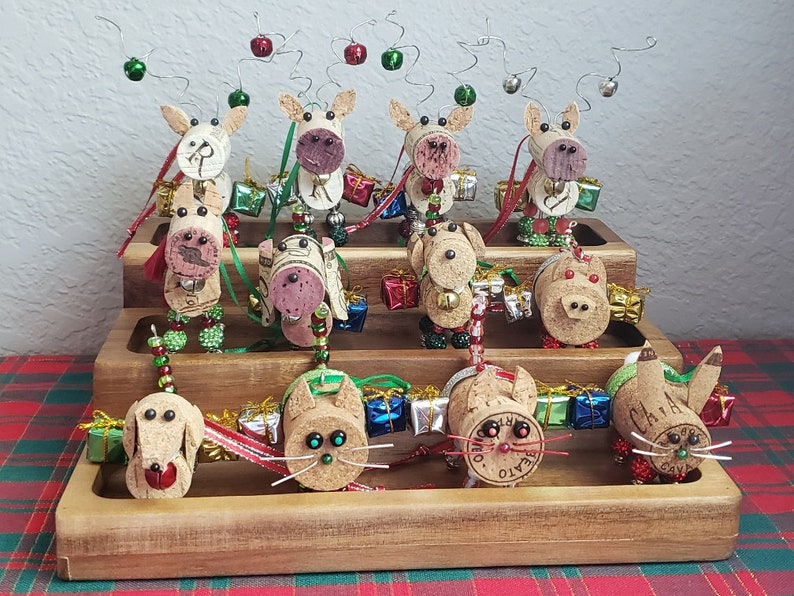May include: A tiered wooden display stand with 12 handmade reindeer and cat ornaments made from wine corks. The ornaments are decorated with beads, bells, and ribbons. The reindeer have red noses and antlers made from wire. The cats have whiskers and tails made from wire.