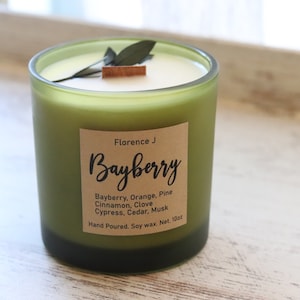 Puede incluir: Un tarro de vela de vidrio verde con una etiqueta marrón que dice "Florence J Bayberry". La etiqueta enumera las notas de aroma: Bayberry, naranja, pino, canela, clavo, ciprés, cedro, almizcle. La vela está hecha a mano y hecha con cera de soja. El tarro tiene un peso neto de 10 oz.