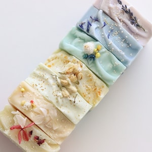 Op de afbeelding: Een set van zeven handgemaakte zeepjes met verschillende kleuren en ingebedde gedroogde bloemen. De zeepjes zijn in een rij gerangschikt, met de lichtste kleur onderaan en de donkerste kleur bovenaan. De kleuren variëren van wit tot lichtblauw, lichtgroen, geel en lavendel.