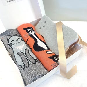 May include: A white gift box contains cat-themed items. Inside are two rolled-up patterned towels, one gray and one orange, and a gray cat-shaped sponge. The box is tied with a gold ribbon. The label reads "OSAEK" and "100% Cotton."