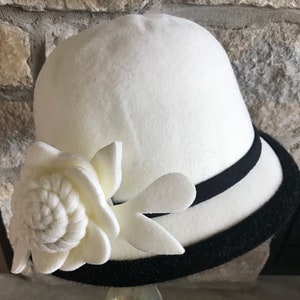 以下が含まれることがあります： 黒いフェルトのバンドと白いフェルトの花のアクセントが付いた、白いフェルトのクロッシェハットです。