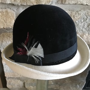 May include: A black velvet hat with a white brim and a black ribbon. The hat is decorated with a small cluster of feathers in shades of red, white, and black.