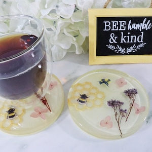 May include: Two round, clear resin coasters with a bee and honeycomb design. The coasters have pressed flowers and glitter embedded in the resin. The coasters are sitting on a white surface. A sign reads "BEE humble & kind".