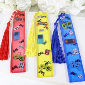 May include: Three rectangular resin bookmarks in red, yellow, and blue, each featuring a matching colored tassel. The bookmarks are decorated with various miniature gaming controller and handheld console graphics scattered across the surface.
