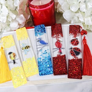 May include: Five colorful bookmarks with different designs. The bookmarks are made of clear resin with glitter and feature a bluebird, a cardinal, a bee, and two ladybugs. The bookmarks are attached to tassels.