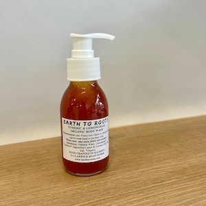May include: A clear bottle with a pump dispenser filled with a bright orange liquid. The bottle label reads "EARTH TO ROOTS TURMERIC & LEMONGRASS ORGANIC BODY WASH" and includes instructions for use and ingredients. The bottle is hand-crafted in Hackney, London.