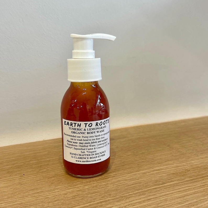 May include: A clear bottle with a pump dispenser filled with a bright orange liquid. The bottle label reads "EARTH TO ROOTS TURMERIC & LEMONGRASS ORGANIC BODY WASH" and includes instructions for use and ingredients. The bottle is hand-crafted in Hackney, London.