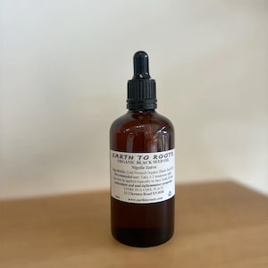 Könnte beinhalten: Eine braune Glasflasche mit Earth to Roots Organic Black Seed Oil und einer schwarzen Pipette. Das Etikett enthält den Text "Nigella Sativa" und Gebrauchsanweisungen. Die Flasche steht auf einer hellbraunen Oberfläche.