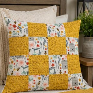 May include: A square throw pillow featuring a patchwork design of alternating mustard yellow honeycomb patterned fabric squares and white fabric squares printed with a pattern of grey beehives, small yellow bees, and colorful wildflowers.