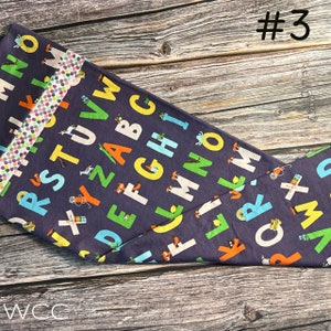 May include: A dark blue fabric with a white, yellow, green, and orange alphabet print. The fabric has a white and pink polka dot trim. The fabric is folded in half and the number #3 is written in white on a black background in the top right corner of the image.
